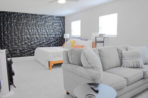 Ein modernes Schlafzimmer mit einem Kopfende im geometrischen Muster, einem grauen Sofa, einem Bett mit vielen Kissen und einem kleinen runden Tisch mit einer Fernbedienung.