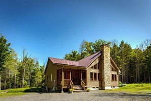 A log cabin with a porch, a stone chimney, and a forested backdrop.