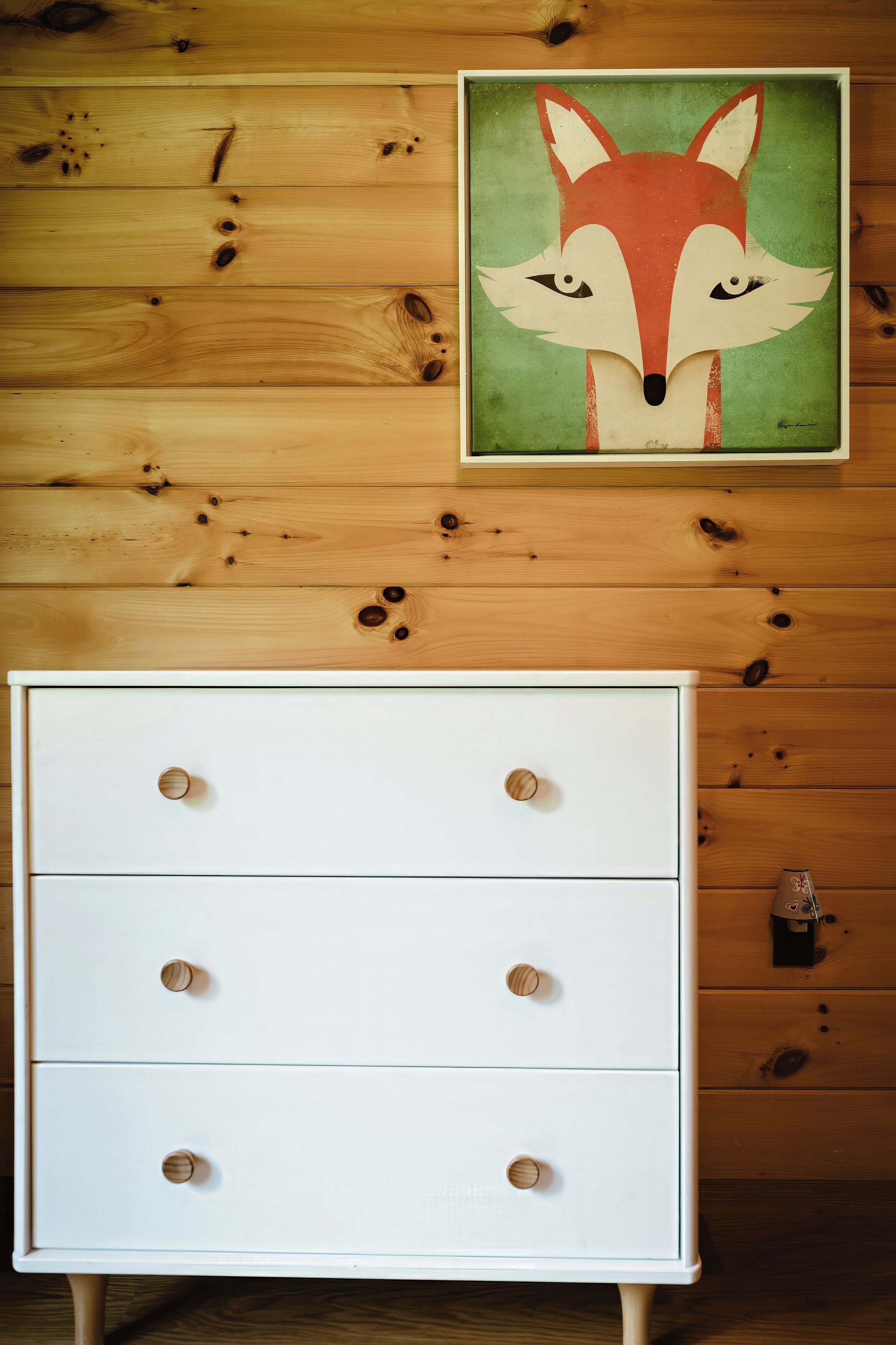 Une commode blanche avec des boutons ronds, placée contre un mur en bois orné d’un tableau représentant un renard.
