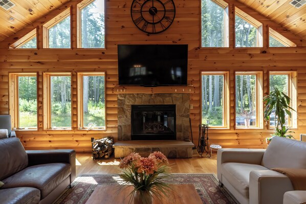 A log cabin interior with a fireplace, a flat-screen TV, and multiple windows offering a forest view.