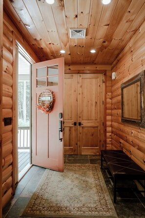 A wooden cabin interior with a pink door, a welcome sign, a bench, and a mirror.