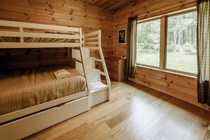 Quarto de cabana de madeira com beliche, escada, janela com cortinas e um pequeno quadro na parede.