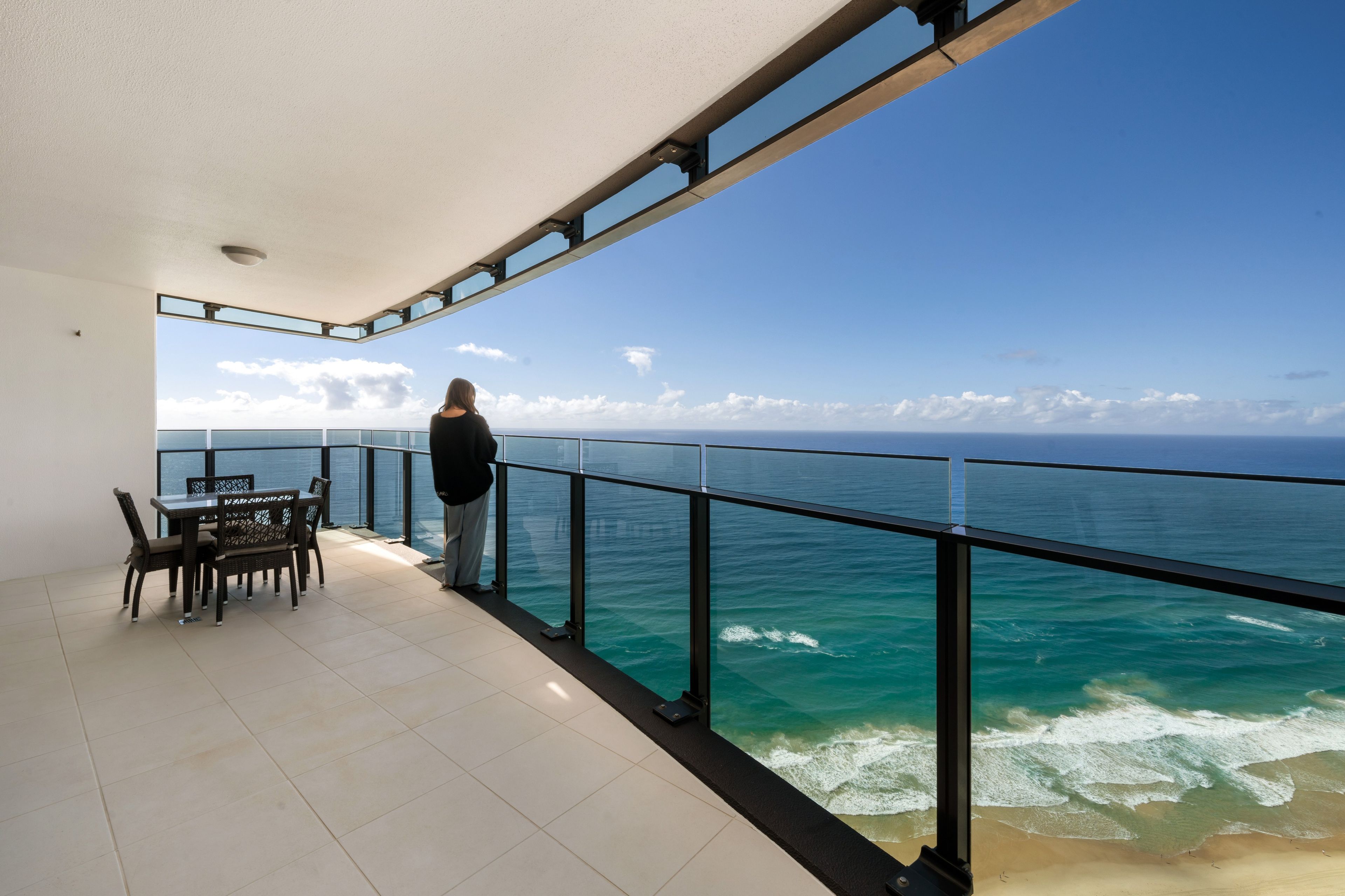 A balcony with a view of the ocean, a table with chairs, and a person standing.