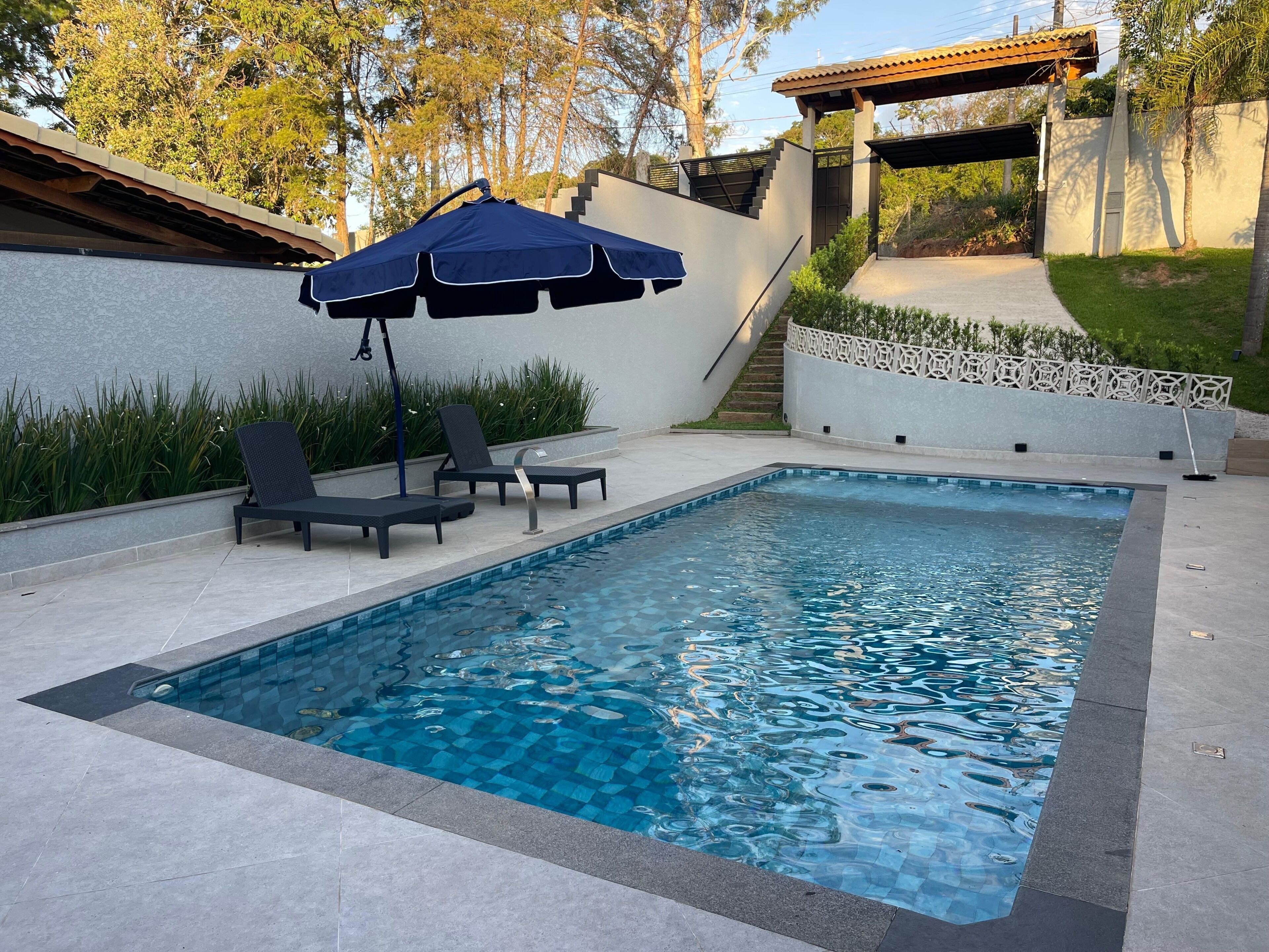 A rectangular swimming pool with a tiled edge, surrounded by a low wall and a covered seating area with a blue umbrella.
