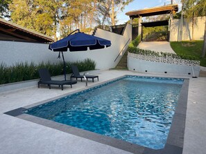 A rectangular swimming pool with a tiled edge, surrounded by a low wall and a covered seating area with a blue umbrella.