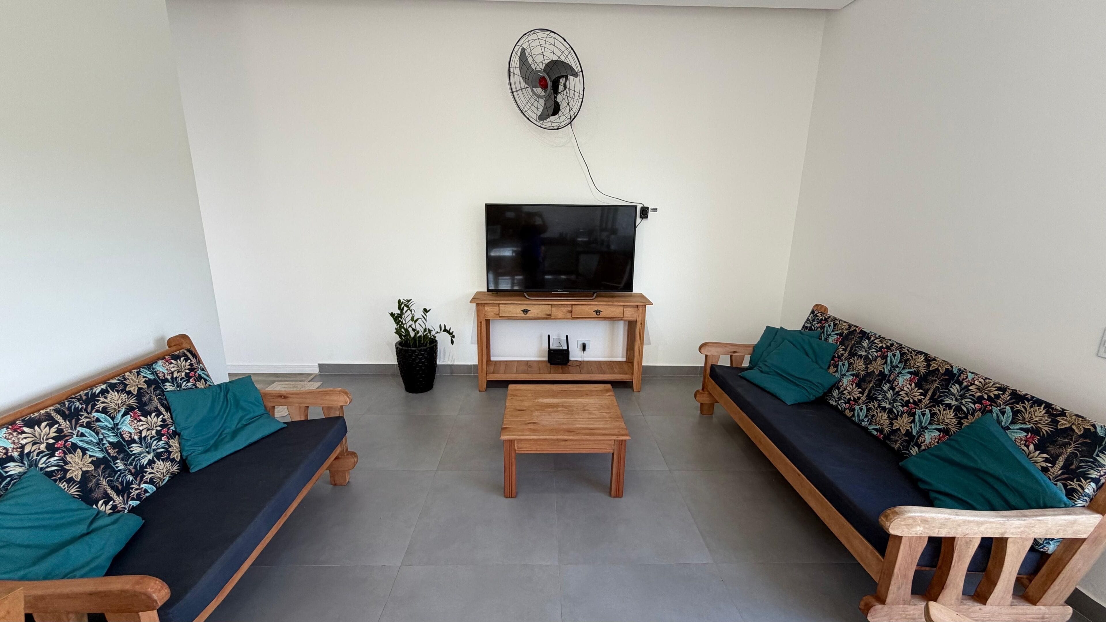 Una sala de estar con dos sofás, una mesa de centro, un televisor sobre un mueble de madera, un ventilador de pared y una planta en maceta.