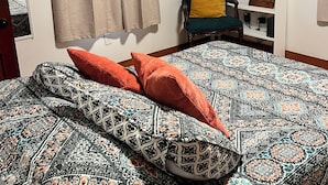 A bedroom with a patterned bedspread, a chair with a cushion, a shelf with various items, and a window with curtains. - Charming 2-bedroom house in peaceful Cunningham (Cunningham)
