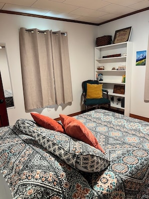 Un dormitorio con una colcha estampada, una silla con cojín, una estantería con diversos objetos y una ventana con cortinas.