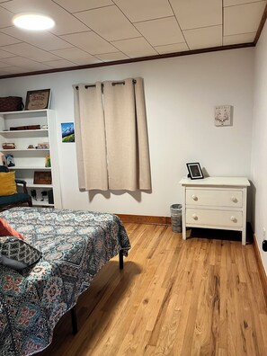 Ein Schlafzimmer mit einem Bett, einem Nachttisch, einem Regal mit verschiedenen Gegenständen, einem Bild an der Wand und einem Fenster mit Vorhängen.