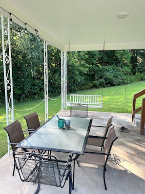 Un patio exterior con mesa y sillas, una hamaca y vistas a un césped verde y árboles.