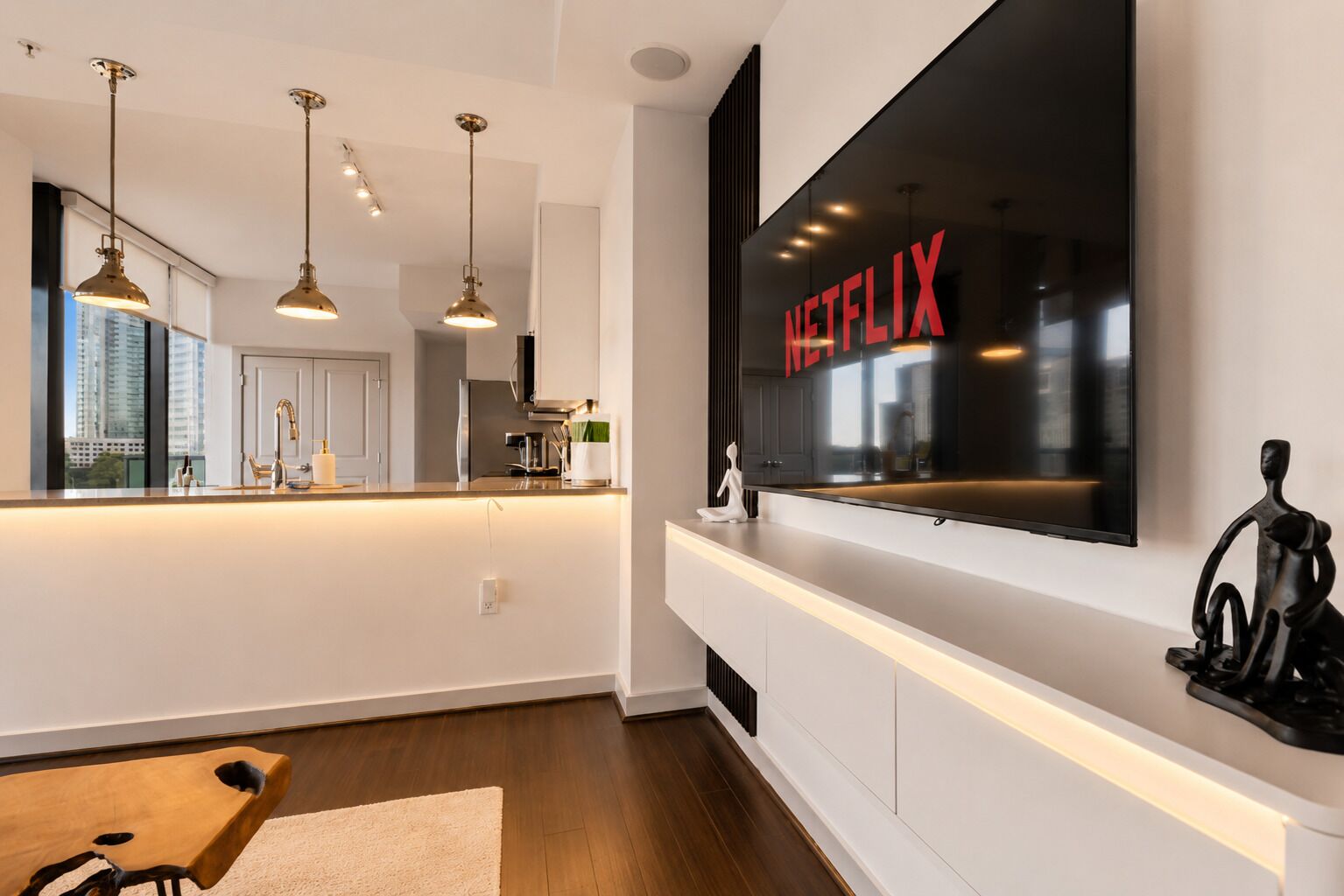 A modern kitchen with pendant lighting, a large flat-screen TV mounted above the counter, and a view of the city through the window.