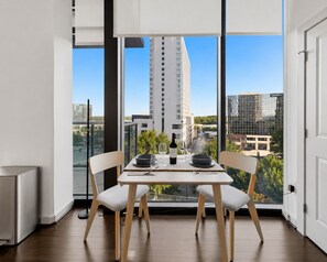 A dining area with a table set for two, large windows with a city view, and a modern building in the background. - Buckhead Luxury 1BR - Lenox City View - Free Parking (Atlanta)