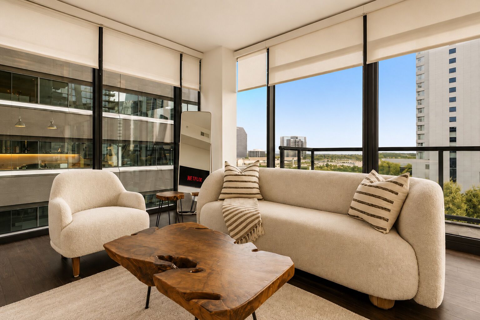 Un salon moderne avec un canapé, un fauteuil et une table basse, de grandes fenêtres offrant une vue sur la ville et un logo Netflix.