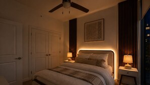 Ein Schlafzimmer mit einem Bett, zwei Nachttischen mit Lampen, einem Deckenventilator, einer Tür und einem gerahmten Bild an der Wand.