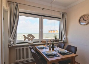 Ein Essbereich mit Blick auf den Strand, ein Holztisch gedeckt mit Geschirr und Besteck sowie eine Wanduhr.