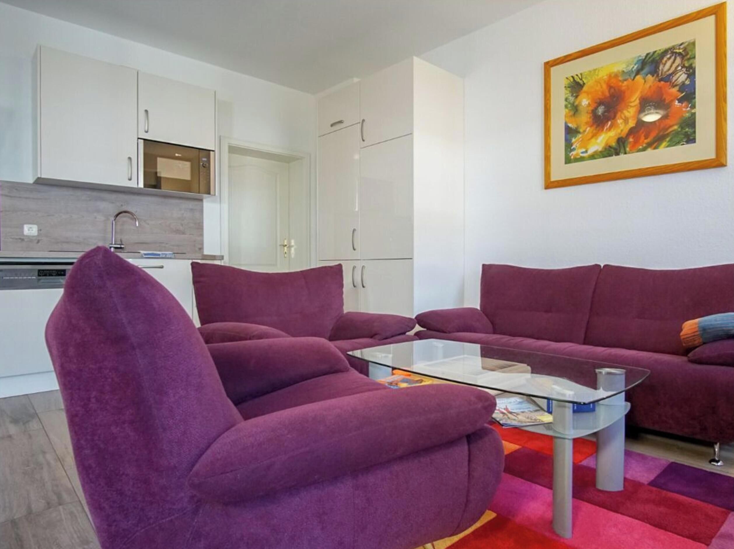 Ein modernes Wohnzimmer mit violetten Sofas, einem Glastisch und einer Küche im Hintergrund.