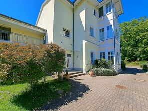 A multi-story building with a paved courtyard, a small garden area with shrubs, and a clear blue sky. - Villa Waldstrasse 03 (Heringsdorf)