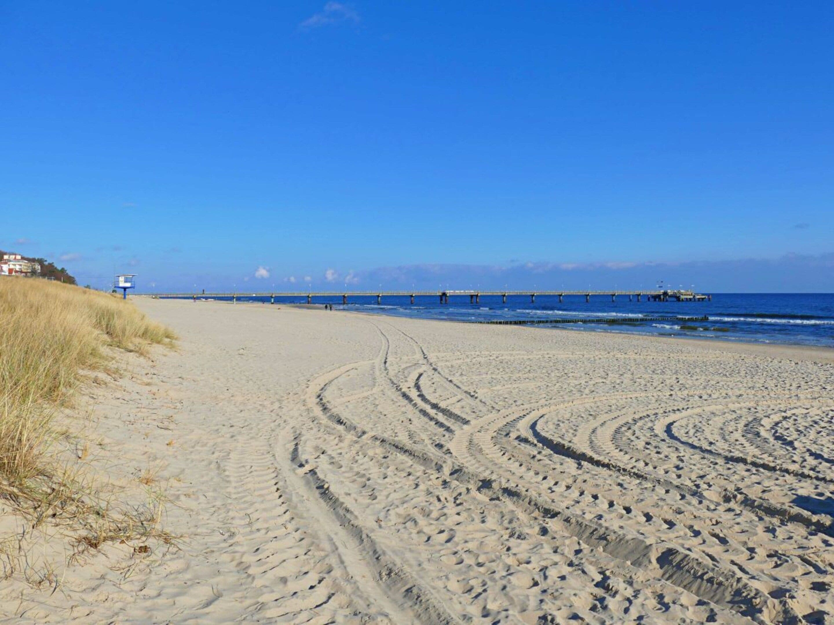 Une plage de sable où l’on voit des traces de pneus menant à l’eau, une jetée au loin et un poste de surveillance des sauveteurs sur la gauche.