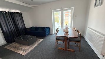 Ein modernes Wohnzimmer mit einem dunkelblauen Sofa, einem Holzespeisetisch mit Stühlen, einem schwarzen Hocker und einem gemusterten Teppich.