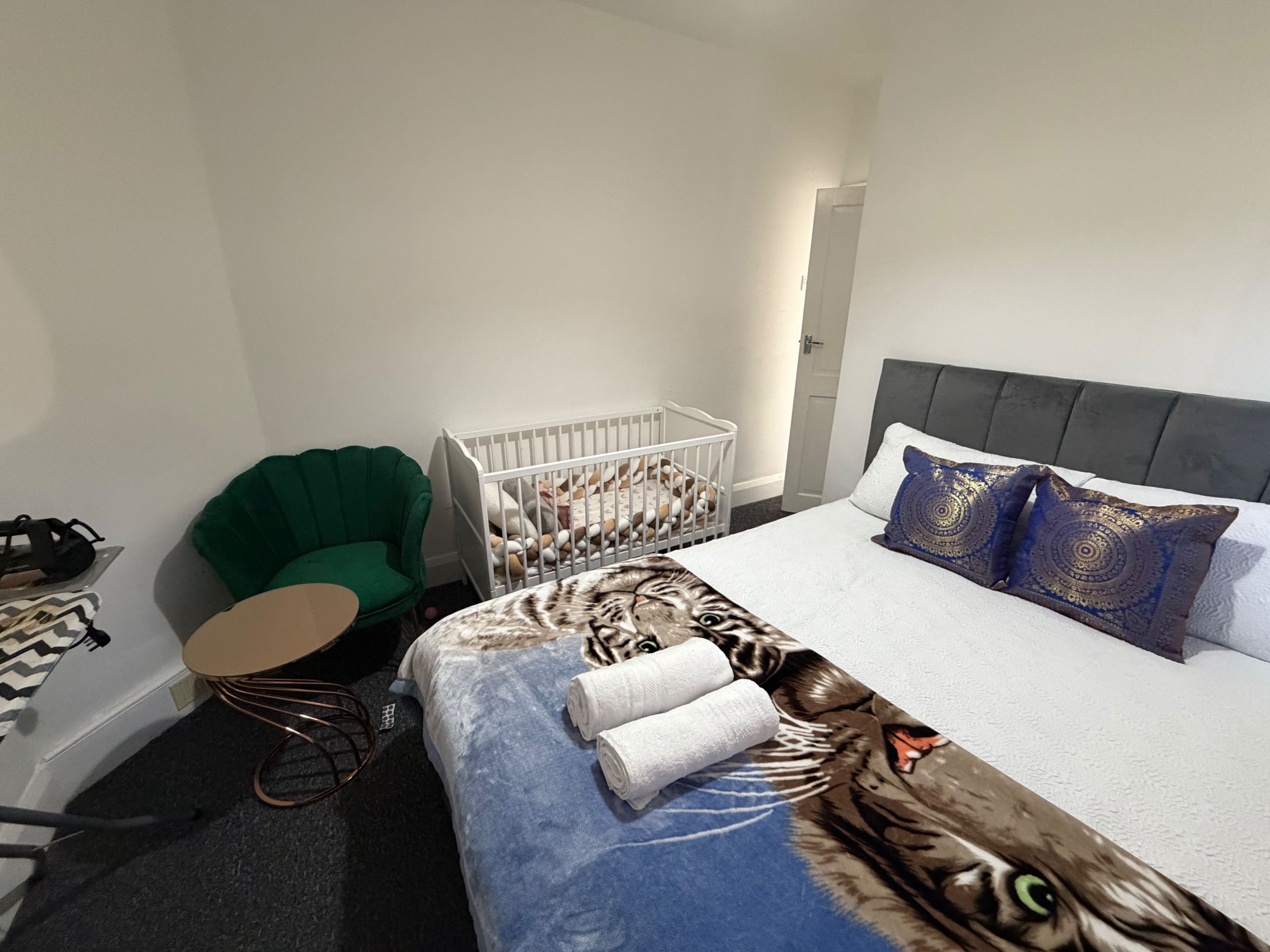 A hotel room with a bed, a green armchair, a white crib, and a small table with a chair.