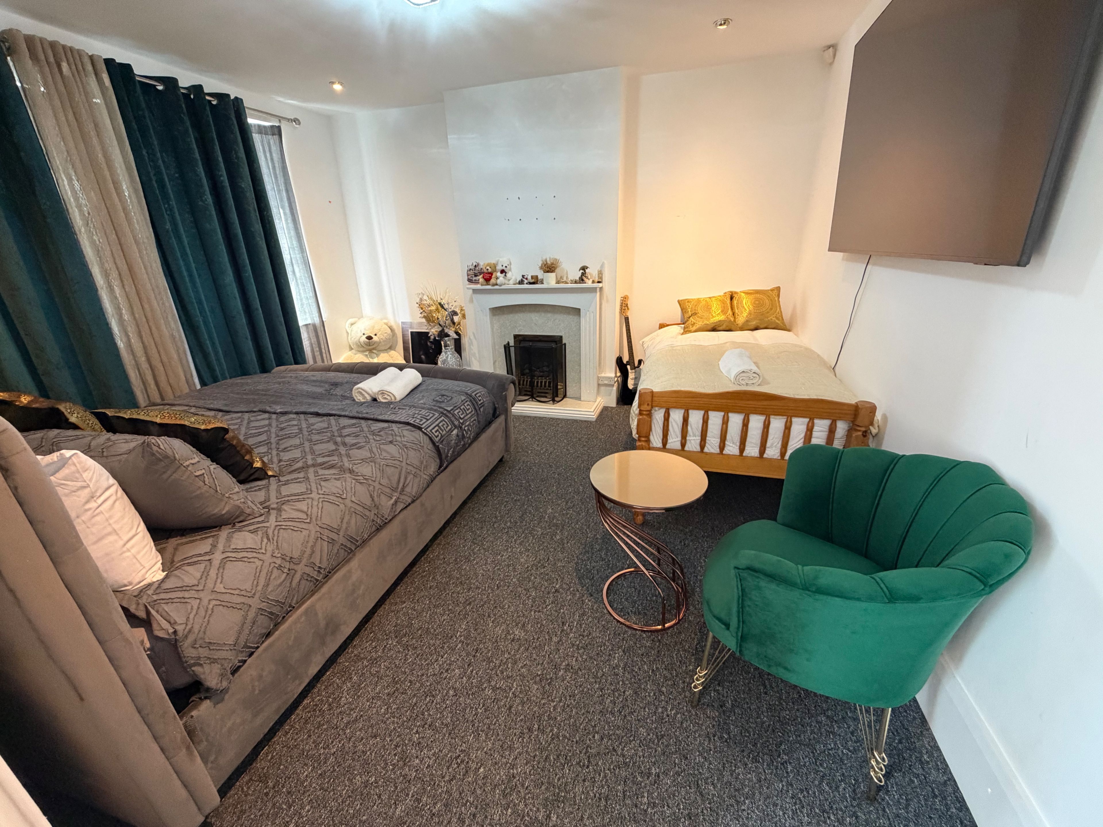 A hotel room with a large bed, a smaller bed, a green armchair, a round side table, and a fireplace.