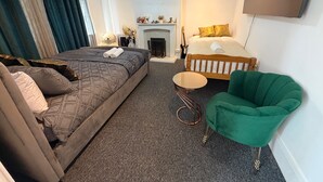 Una habitación de hotel con una cama grande, una cama más pequeña, un sillón verde, una mesita redonda y una chimenea.