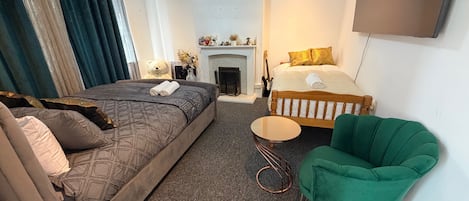 Une chambre d’hôtel avec un grand lit, un lit plus petit, un fauteuil vert, une petite table ronde et une cheminée.