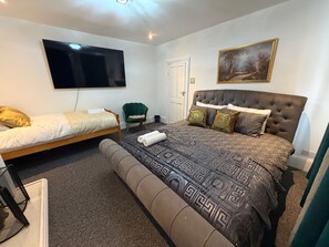 Ein Schlafzimmer mit einem großen Bett, einem kleineren Bett, einem Fernseher, einem Sessel und einem Bild an der Wand.