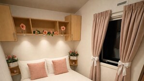 Una cama bien hecha con un detalle en rosa, una ventana con cortinas y una estantería con flores.
