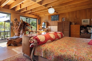 Ein Zimmer mit Holzboden, einem Bett mit floralem Überwurf, einem Holzschrank, einem Spiegel und Blick ins Freie.