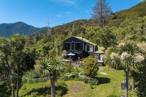 Ein zweistöckiges Haus mit Terrasse, umgeben von üppiger Vegetation und Bergen im Hintergrund.