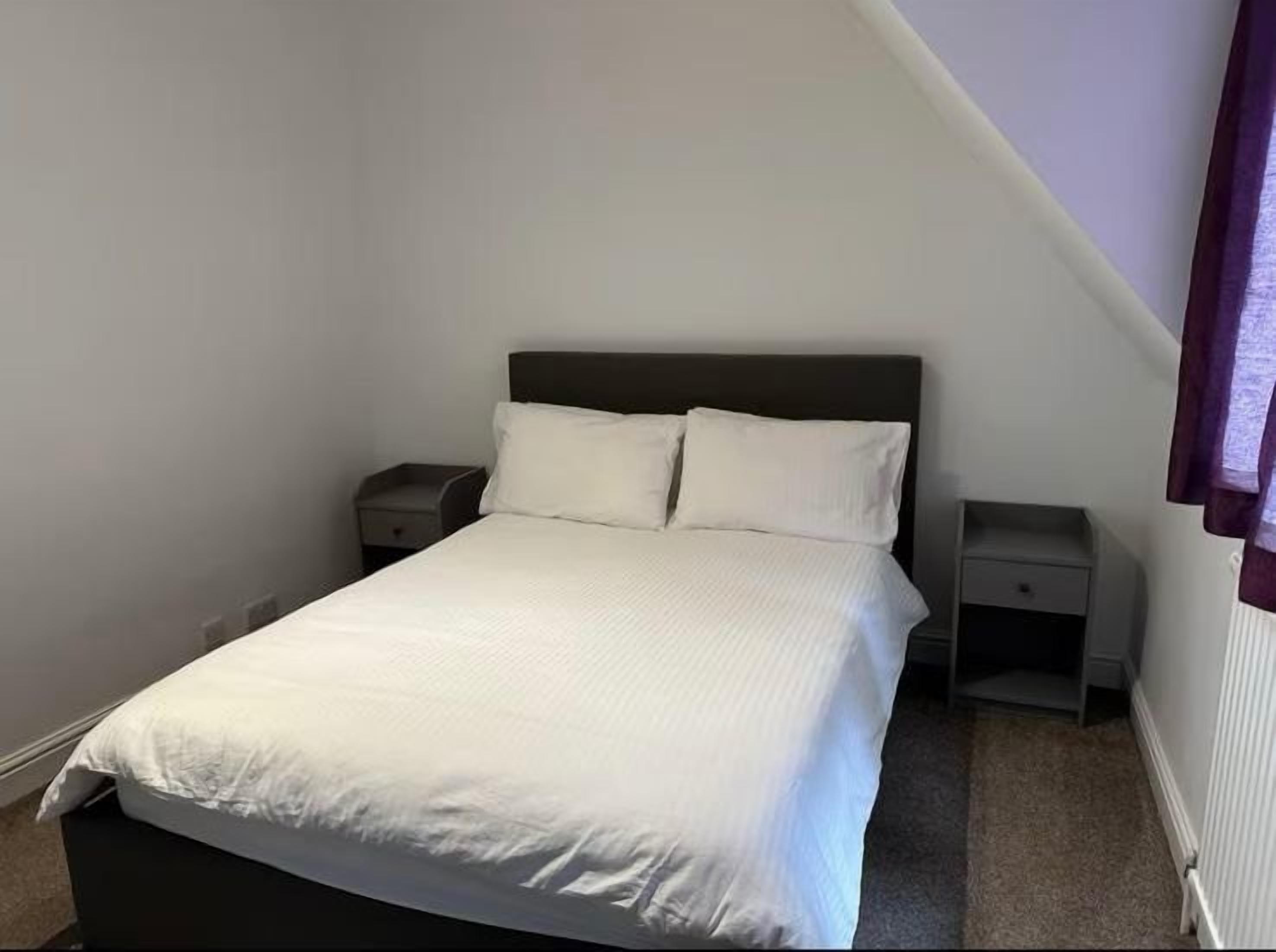 Un lit simple avec une tête de lit sombre, dans une pièce aux murs blancs et au plafond en pente. Il y a deux tables de chevet, une de chaque côté du lit.