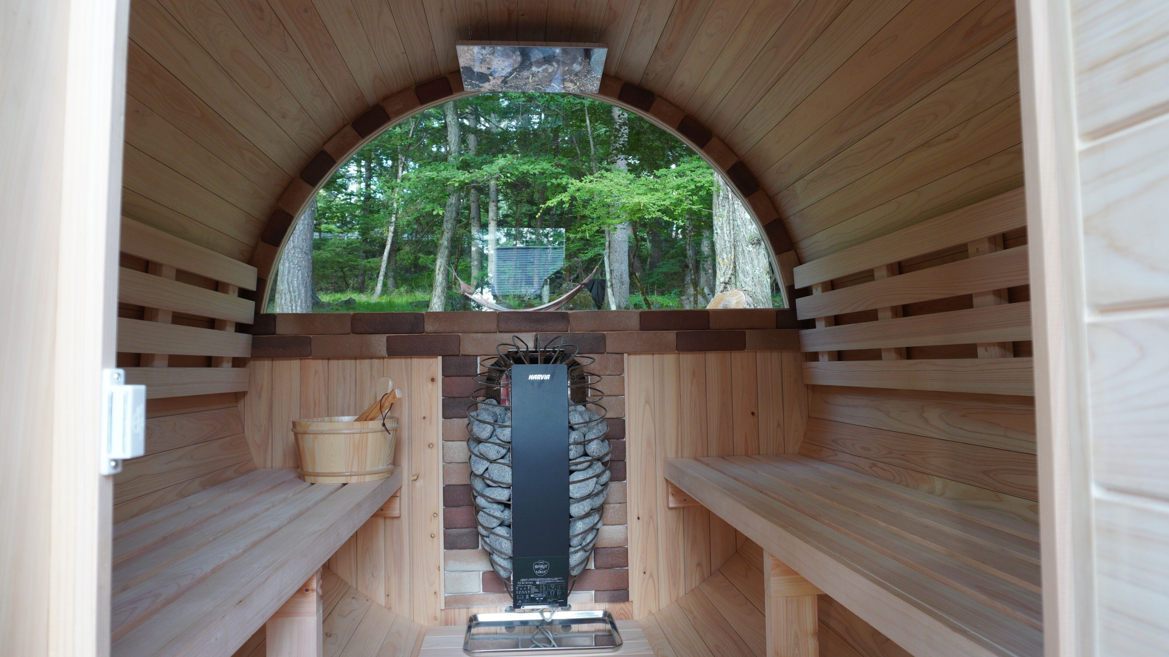 A sauna with wooden benches, a stone wall, and a view of trees through a semi-circular opening.