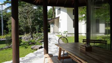 Mesa e banco de madeira em área externa coberta, com vista para uma casa e jardim.