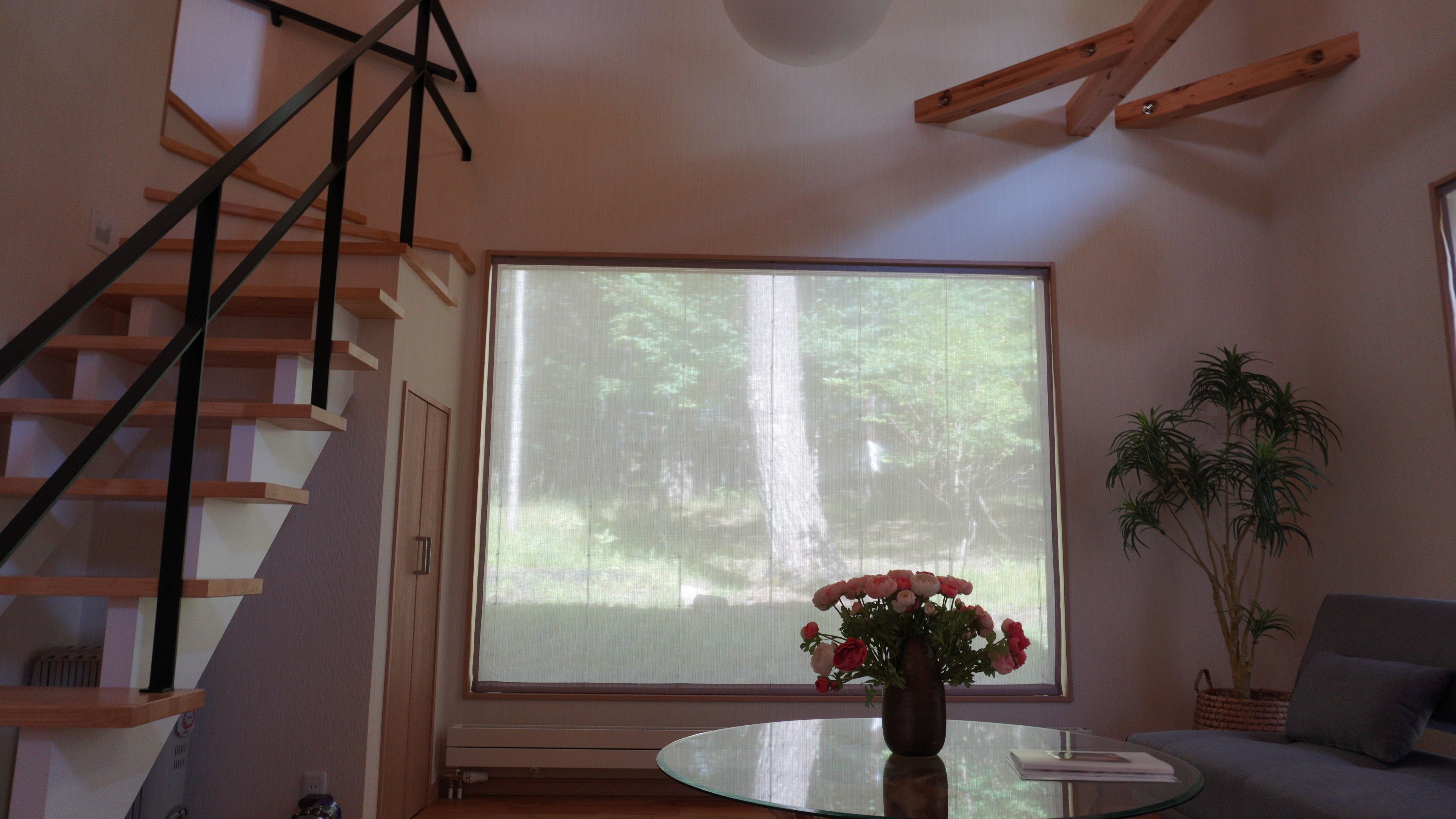 A room with a large window, a staircase, a round glass table with a vase of flowers, and a potted plant.