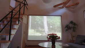 Una habitación con un ventanal grande, una escalera, una mesa redonda de cristal con un florero y una planta en maceta.