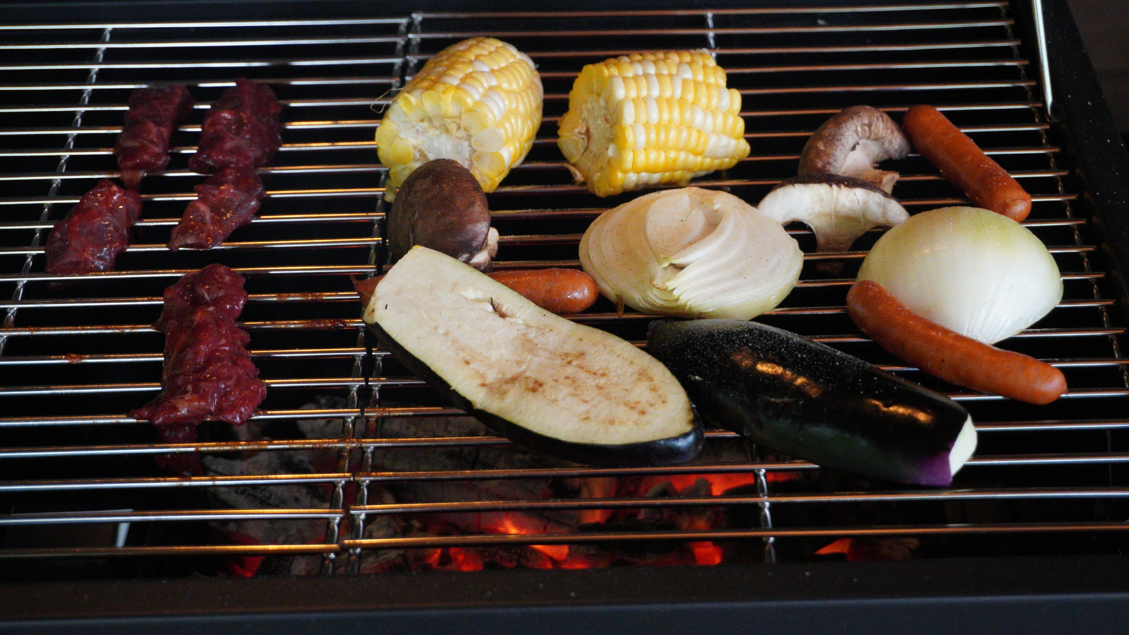 Ein Grill mit verschiedenen zubereiteten Speisen, darunter Maiskolben, Pilze und Würstchen.