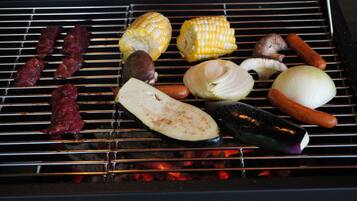 Ein Grill mit verschiedenen zubereiteten Speisen, darunter Maiskolben, Pilze und Würstchen.
