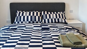 Una cama con colcha a cuadros, dos almohadas, una mesita de noche blanca y una toalla doblada.