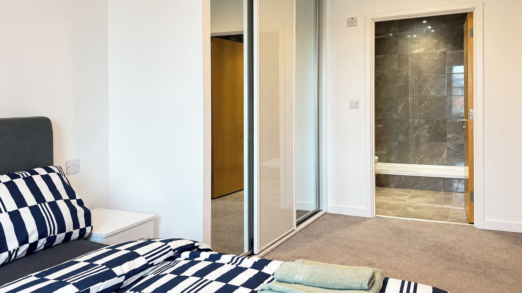Un dormitorio con cama, mesita de noche, baño con ducha y puerta de vidrio que da a un pasillo.