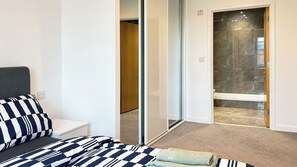 Ein Schlafzimmer mit Bett, Nachttisch, Badezimmer mit Dusche und Glastür zu einem Flur.