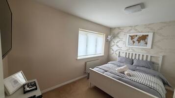 A bedroom with a bed, a window, a radiator, a framed picture on the wall, and a bedside table with a lamp and a remote control.
