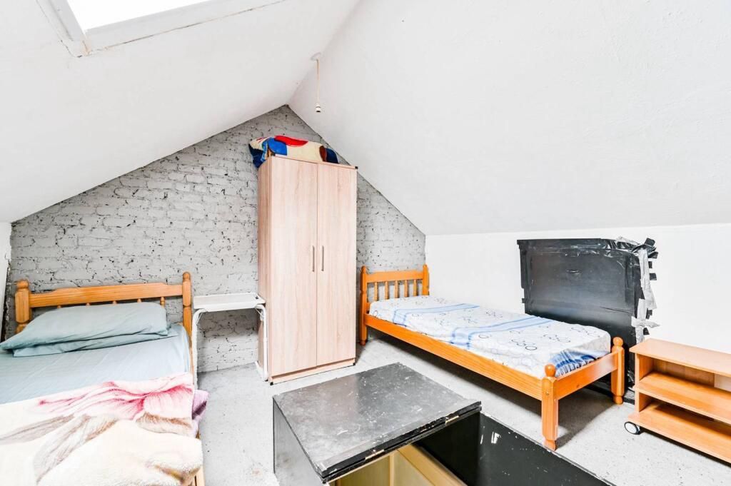 A small, attic-style bedroom with a sloped ceiling, a wooden wardrobe, two single beds, a small table, and a black trash bin.