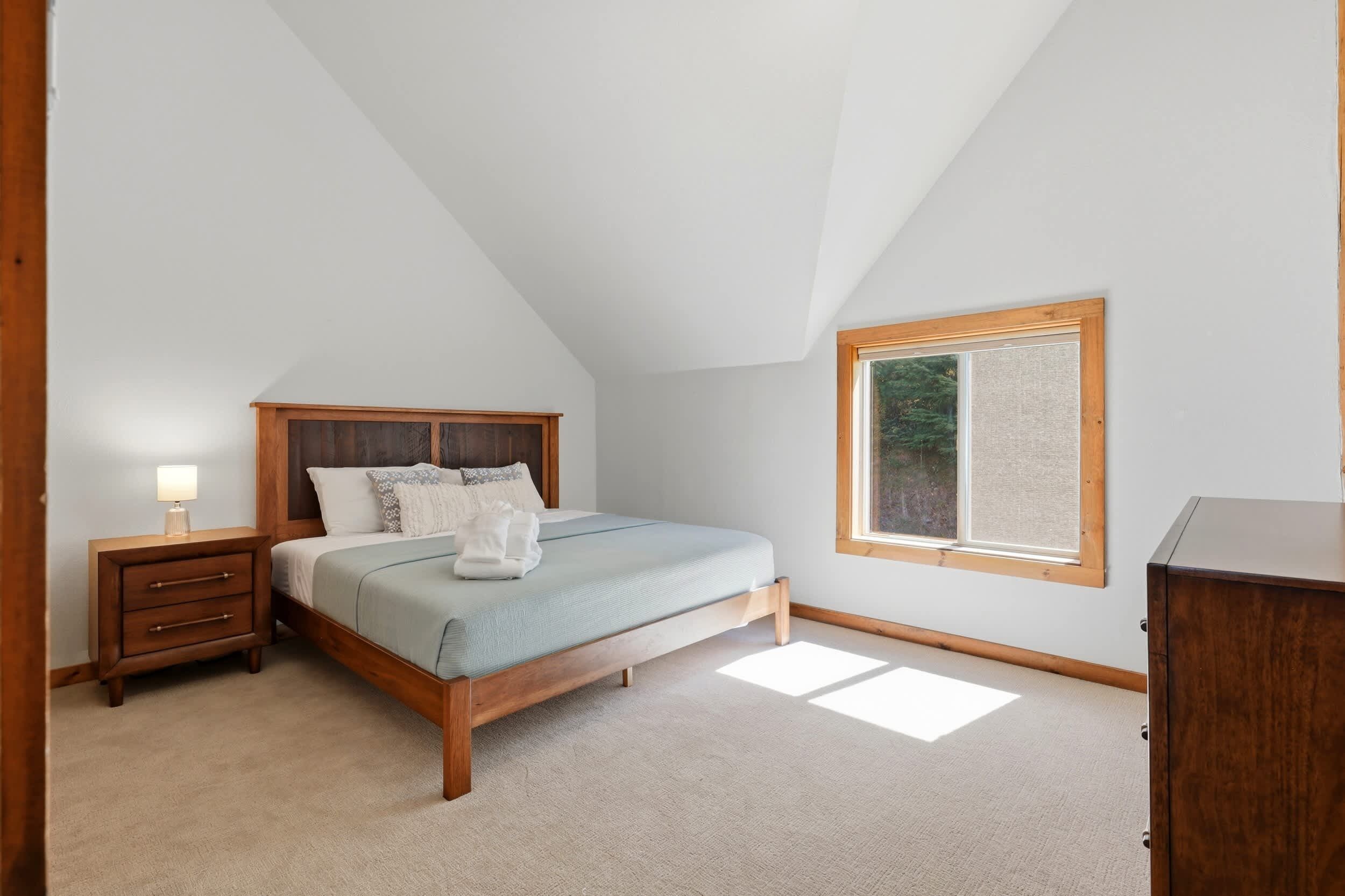 Ein Schlafzimmer mit schräg angesetzter Decke, einem Bett mit Holz-Kopfteil, einem Nachttisch, einer Kommode und einem Fenster mit Blick auf Bäume.