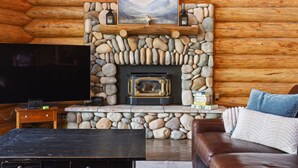 Interior de una cabaña de troncos con chimenea de piedra, un televisor y un sofá de cuero.