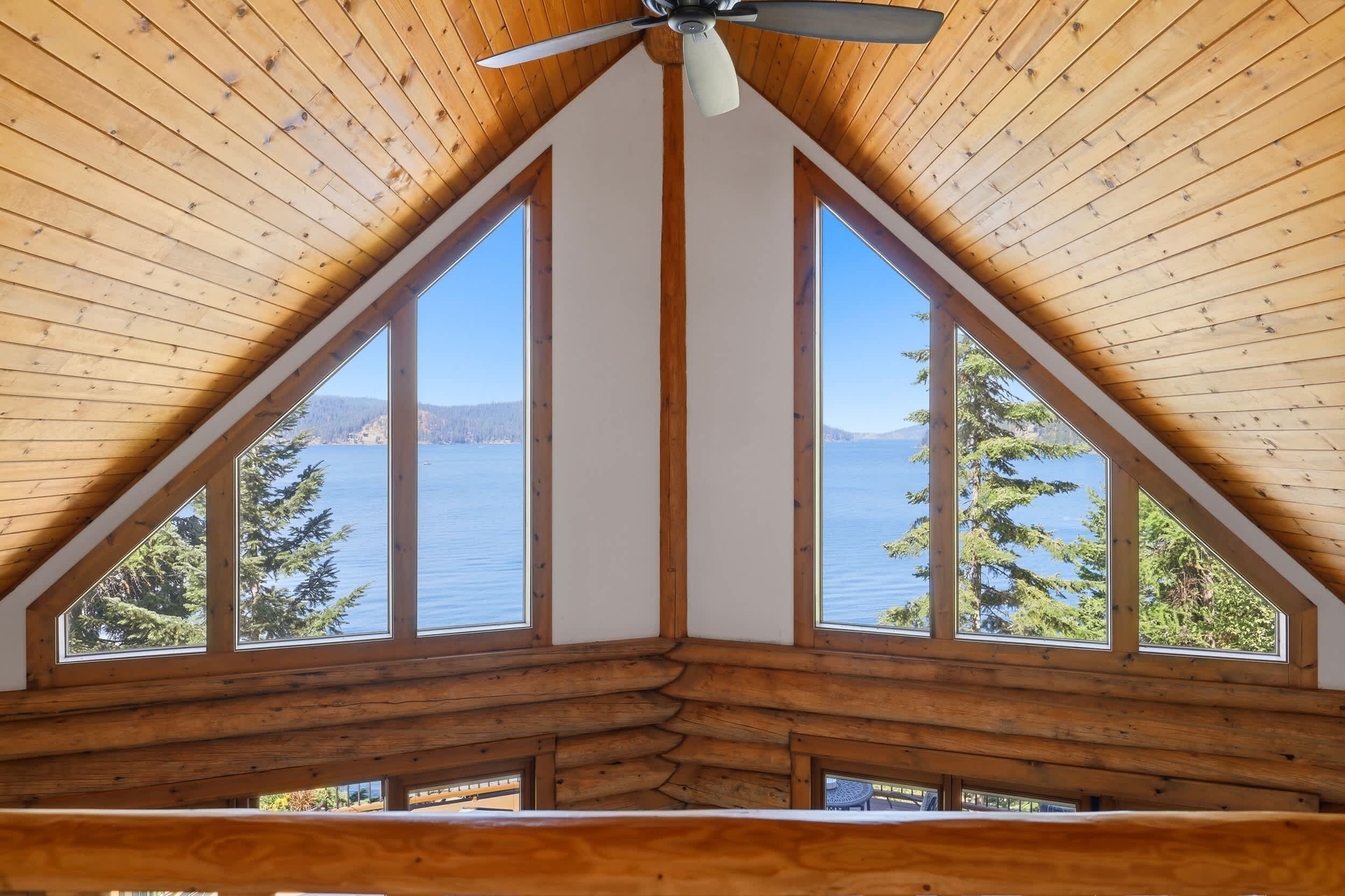 Una cabaña con techo de madera inclinado, ventilador de techo y ventanas amplias con vista a un lago y árboles.