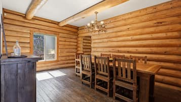 A log cabin interior with a wooden dining table, chairs, a chandelier, and a window.