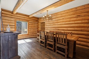 Interior de una cabaña de troncos con una mesa de comedor de madera, sillas, una araña y una ventana.