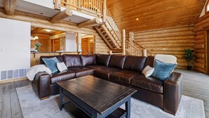 Interior de una cabaña de troncos con un amplio sofá de cuero en forma de L, una mesa de centro de madera y una escalera que sube a un nivel superior.