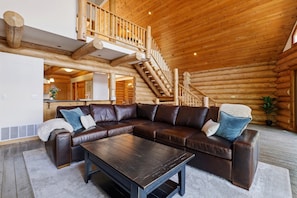 A log cabin interior with a large L-shaped leather sofa, a wooden coffee table, and a staircase leading to an upper level. - Cozy's Lakefront Lodge | Luxury Log Cabin (Harrison)